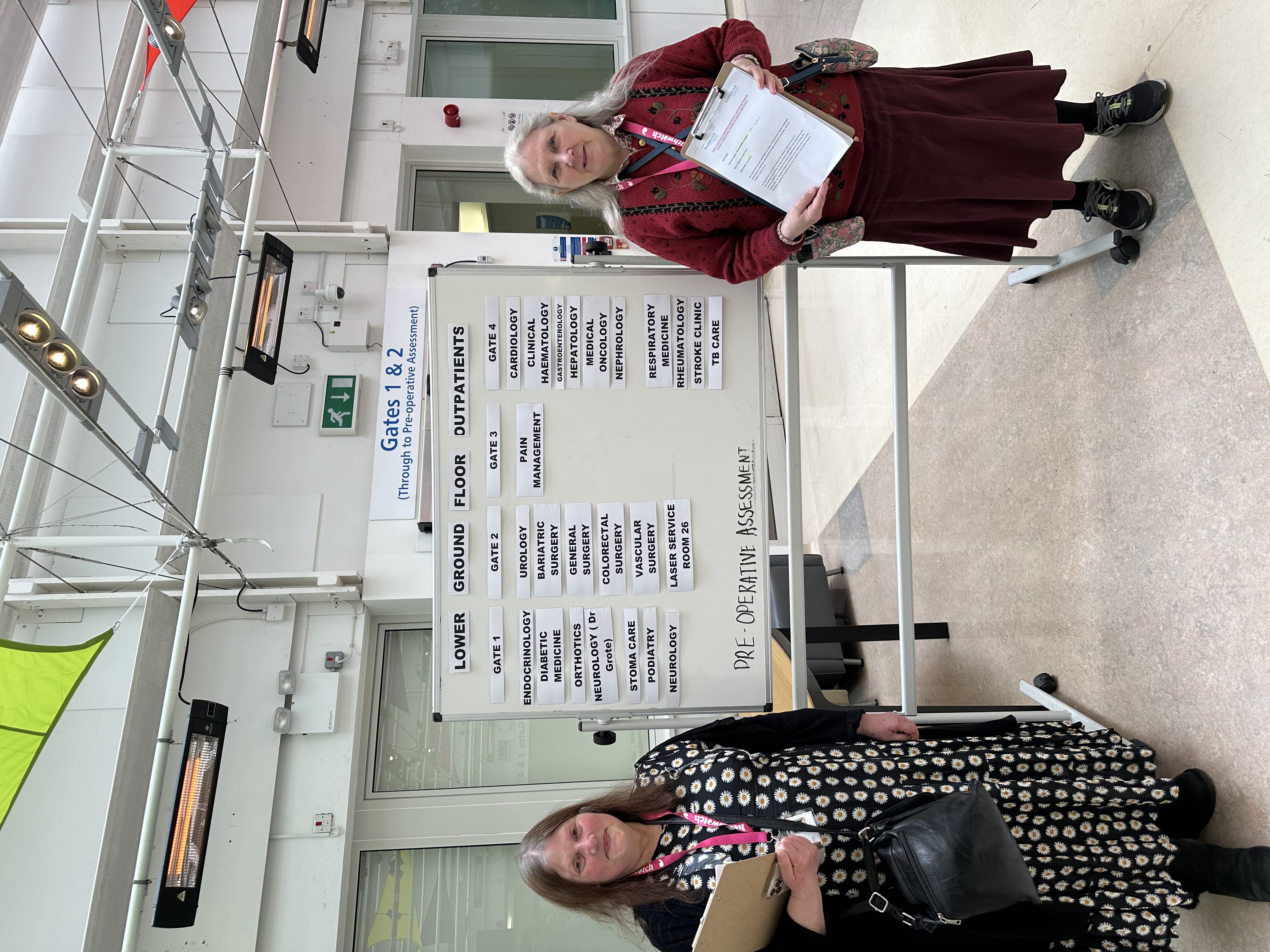 Two volunteers standing next to a board that displays all the outpatient departments that can be found at Chelsea and Westminster Hospital.