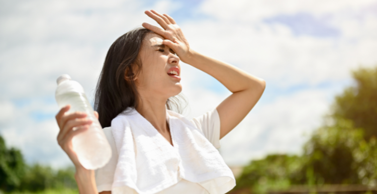 Picture of a lady standing in the sun holding a a bottle of water and putting her hand on her head to stop the sun from getting in her eyes.