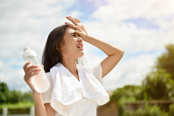 Picture of a lady standing in the sun holding a a bottle of water and putting her hand on her head to stop the sun from getting in her eyes.