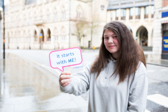 A volunteer holding up a small Healthwatch sign that says "It starts with me"